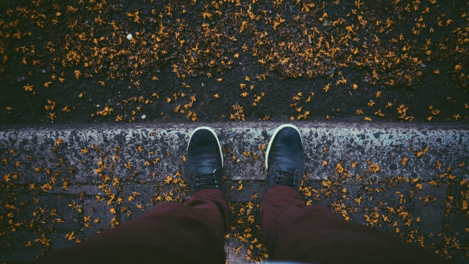 A person's dark shoes beside a kerb scattered with fallen leaves