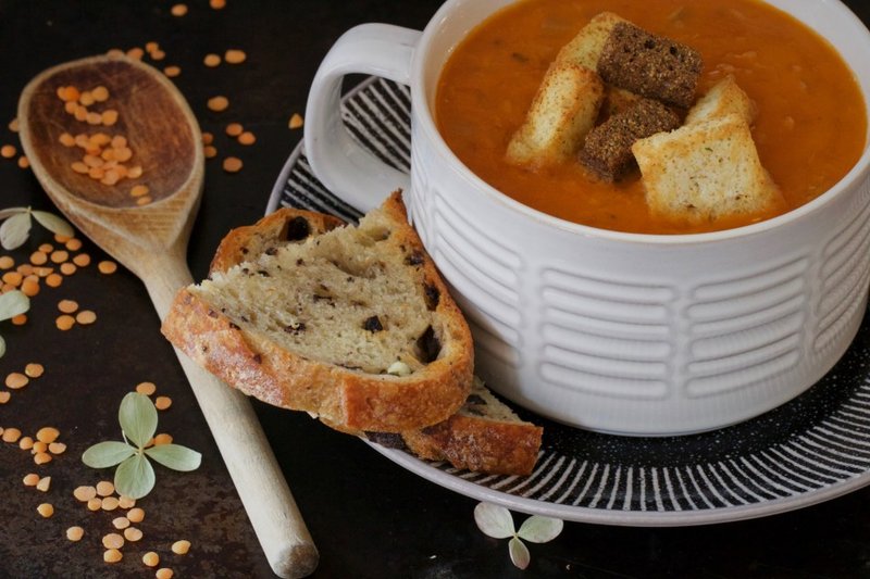 A cup of orange soup with croutons, sliced bread and dried lentils