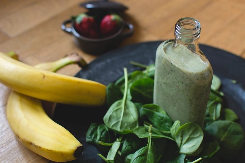 A bottle of green smoothie beside spinach leaves and bananas