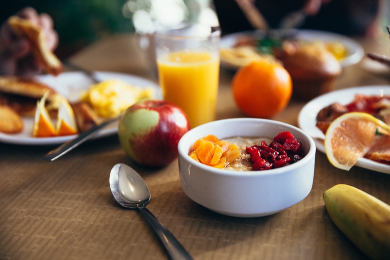 A breakfast table with a bowl of porridge topped with fruit, an apple, a glass of orange juice and plates of food