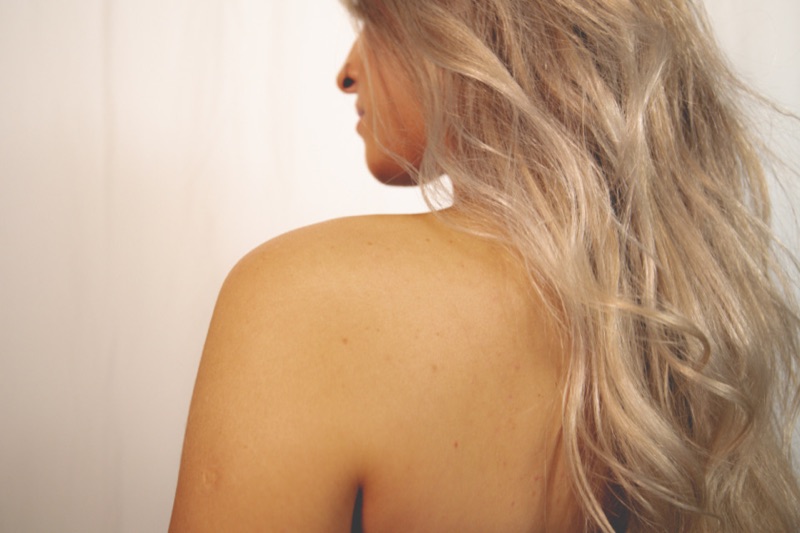 Long, wavy blonde hair falling over a woman's bare shoulder, seen from behind in soft light