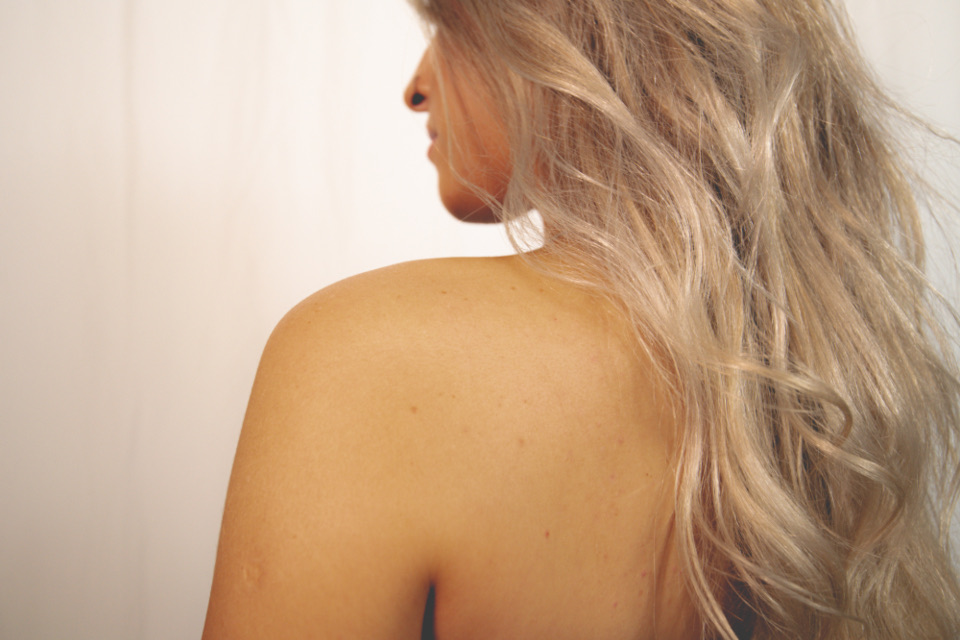 Long, wavy blonde hair falling over a woman's bare shoulder, seen from behind in soft light
