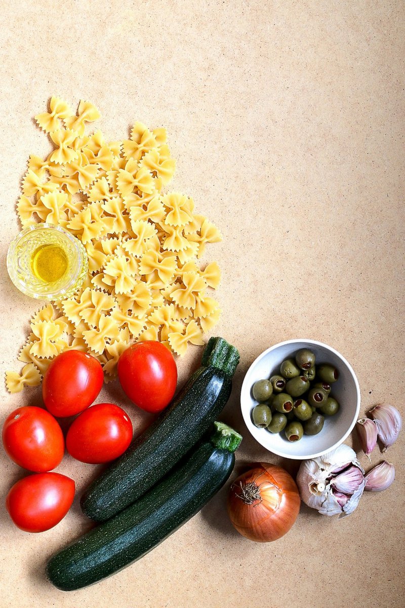 Uncooked pasta, tomatoes, courgettes, olives, onion and garlic arranged on a pale surface