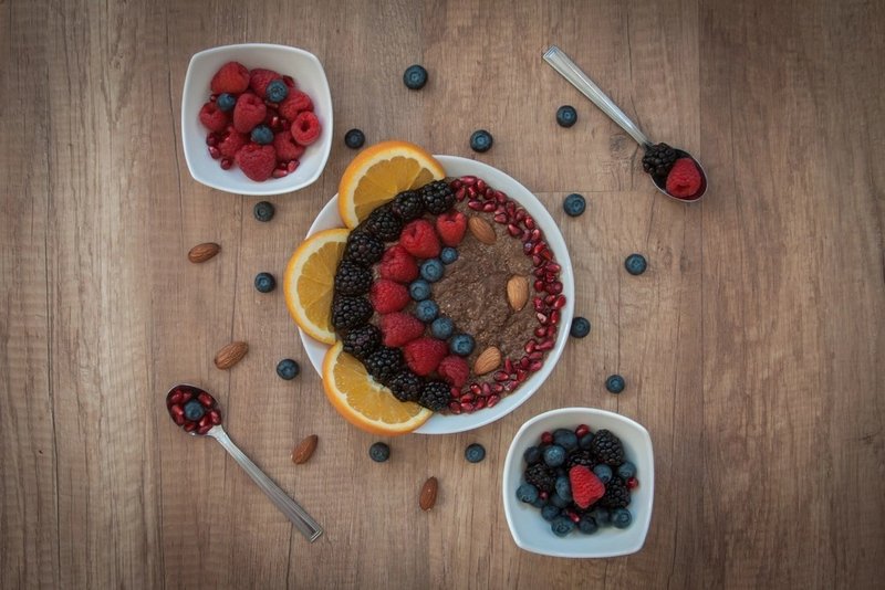 A breakfast bowl topped with berries, almonds and seeds beside sliced orange