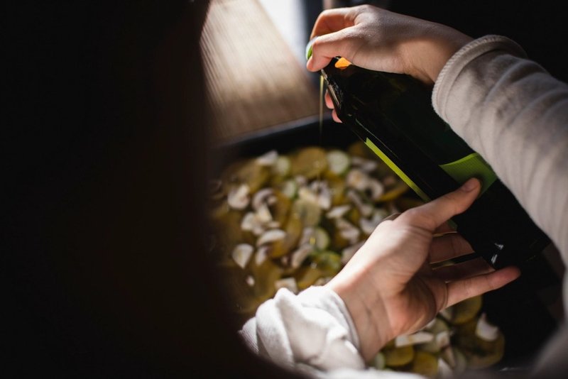 Hands pouring oil from a dark bottle over vegetables in a roasting tray