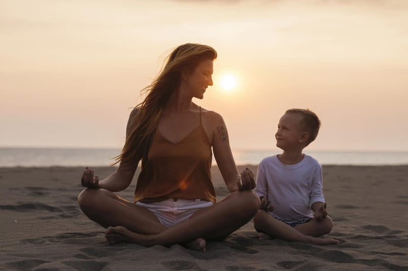 An adult and child sitting cross-legged together on a beach at sunset