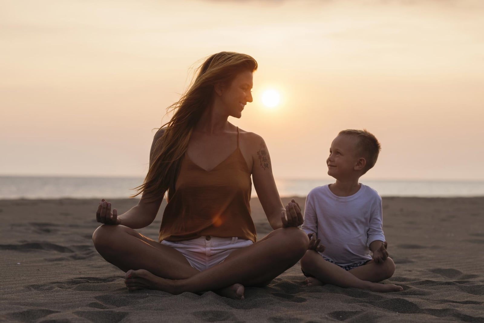An adult and child sitting cross-legged together on a beach at sunset