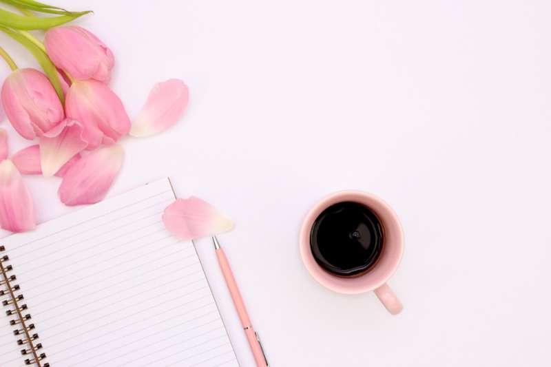 Pink tulips and petals beside an open lined notebook, a pink pen and a cup of black coffee