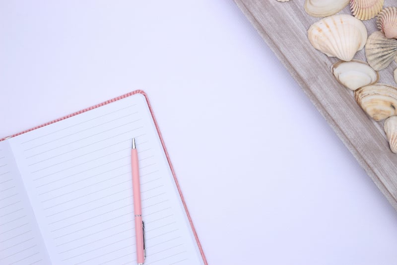 A blank lined notebook with a pink pen beside pale seashells