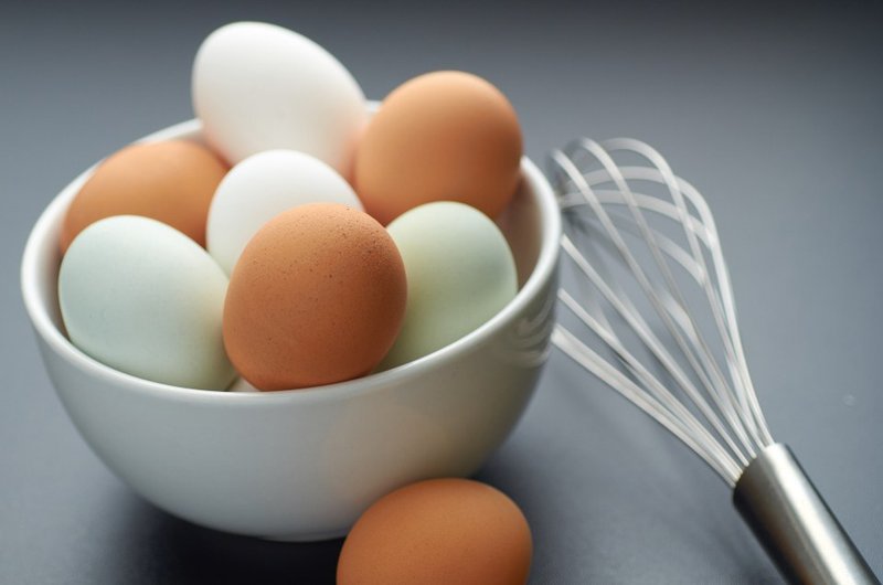A white bowl filled with eggs beside a metal whisk